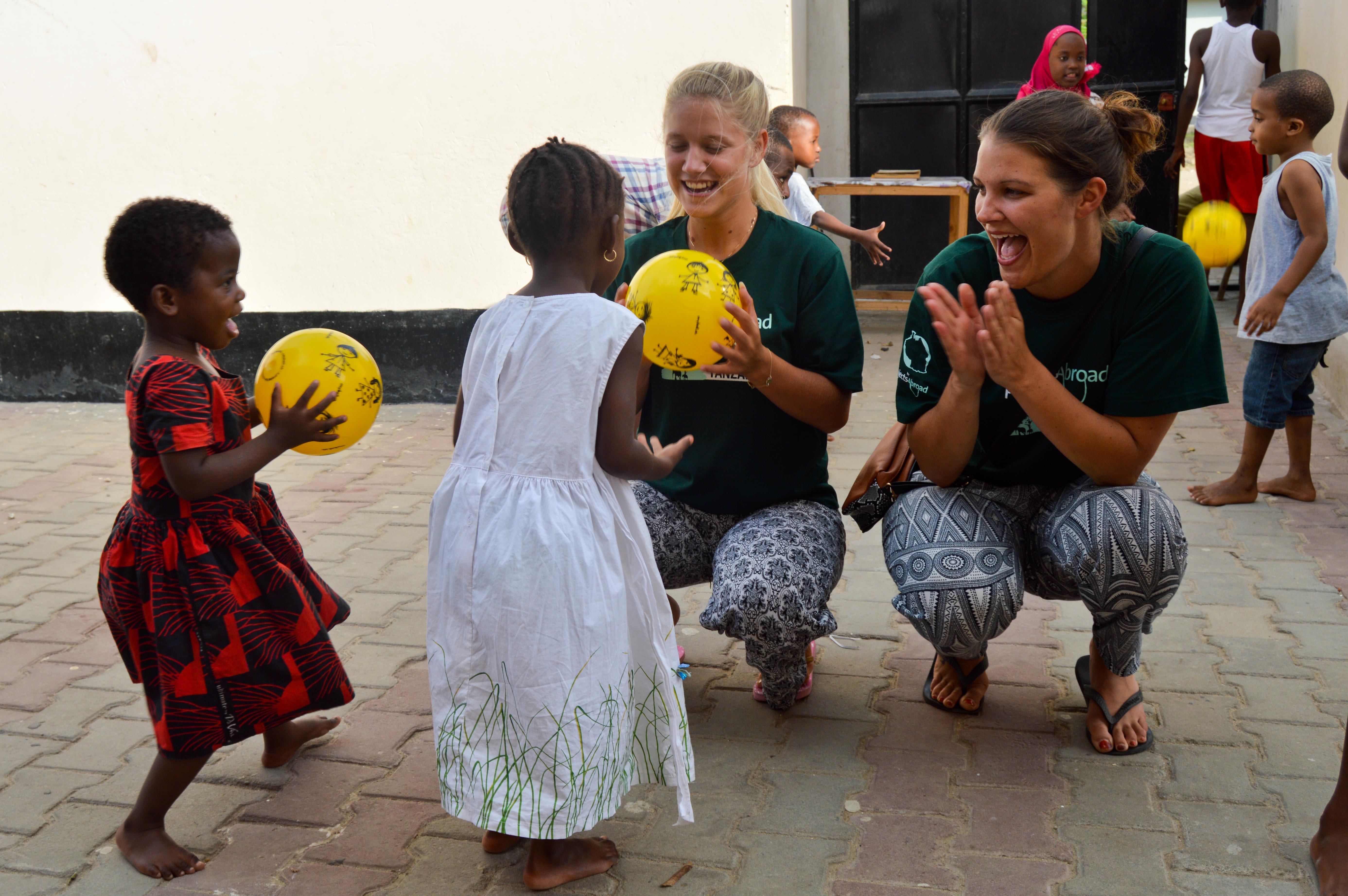 Two female Childcare volunteers play ball games with chilrden outside kindergarten on Care placement. Projects Abroad volunteers with children in Tanzania play ball activities outside a kindergarten.